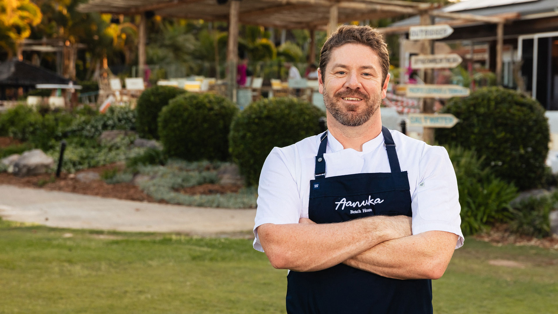 Chef Brad Sloane, in a white jacket and navy apron with “Aanuka Beach House” embroidered, stands outdoors with arms folded, smiling in front of a lush resort backdrop.
