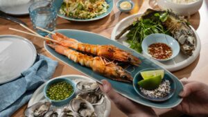 Grilled prawns on skewers served on a blue platter with lime wedges and a dish of salt and pepper for muối tiêu chanh. Surrounding dishes include oysters with cucumber dipping sauce, fried whole fish with herbs and nước chấm, and a fresh salad.