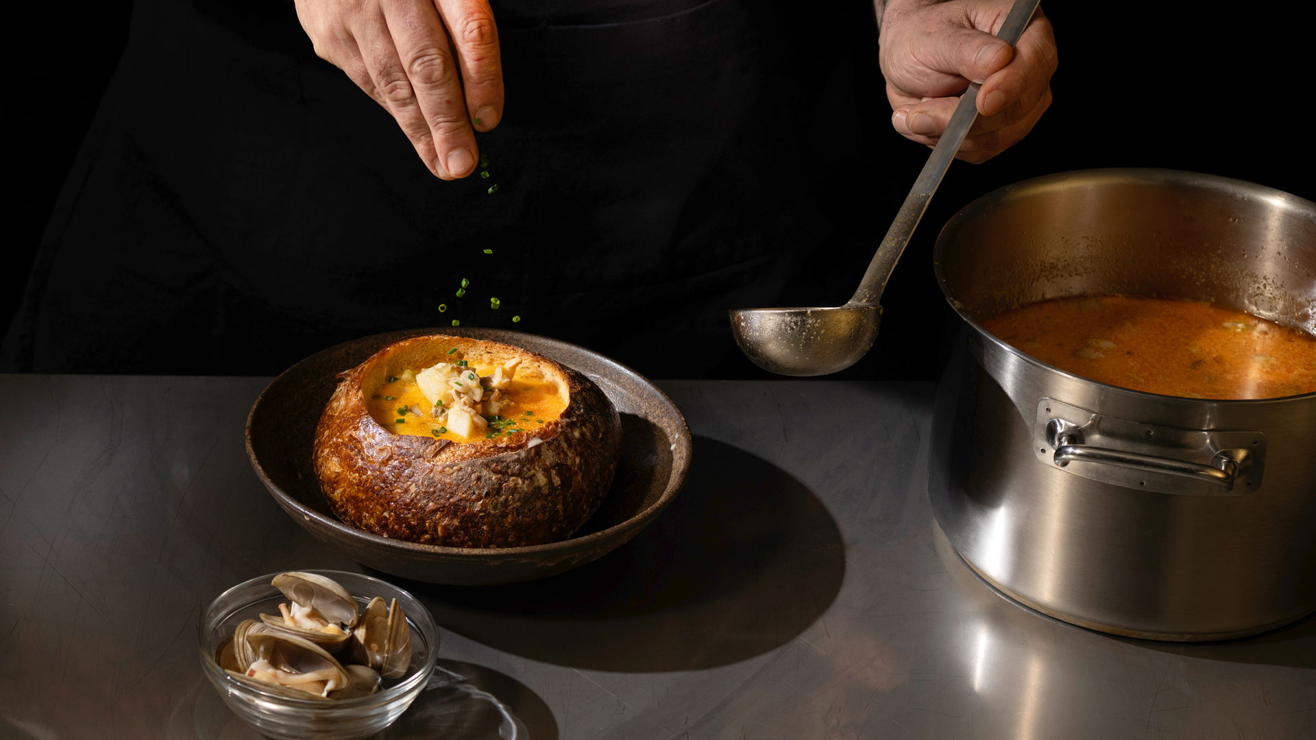 Chef garnishing clam chowder served in a hollowed sourdough bread bowl, with a pot of chowder and a bowl of clams on a stainless steel counter.